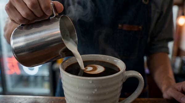 A barista in a small Tokyo café pours steamed milk into a dark espresso from 15cm height in one…