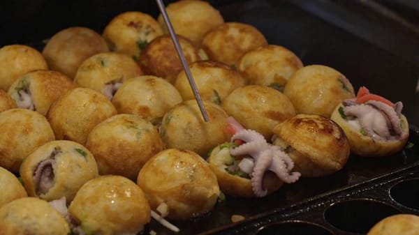 陽気な日本の屋台店主が、熱い鉄板でたこ焼きを長い金属串で器用にひっくり返す。 衣がジュージュー音を立てて焼き色がつき、タコ、紅生姜、ねぎが顔を出す。蒸気が勢いよく立ち上る中、暖かい赤い提灯が…