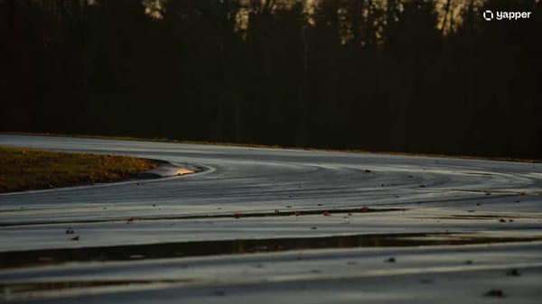 A sleek matte-black sports car enters the frame from the left at high speed on a rain-slicked a…