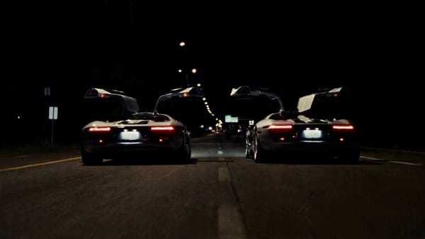 Two sleek silver supercars with glowing red taillights race side by side down a dark nighttime…