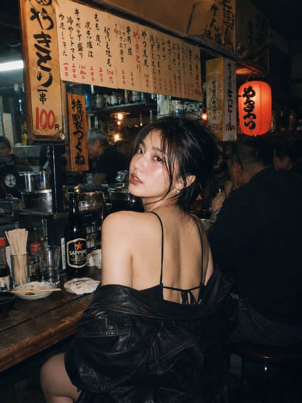 A beautiful young East Asian woman with long dark wavy hair and striking makeup, sitting at a woode… prompt example