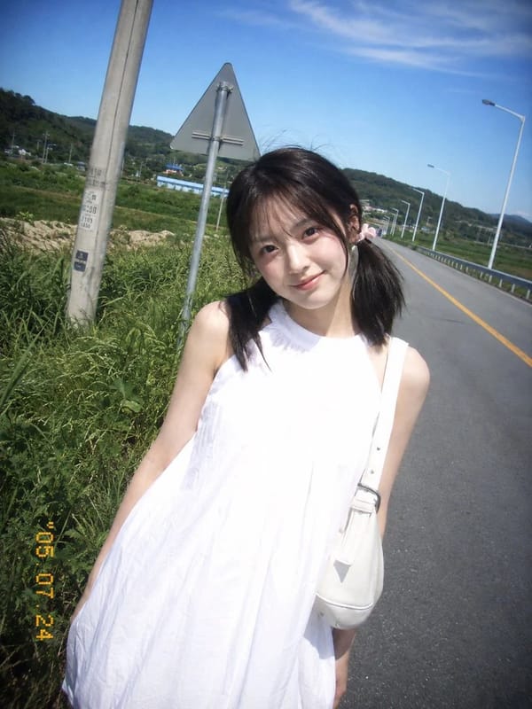 复古 CCD 卡片机夏日旅行抓拍，一位20岁出头的年轻东亚女idol站在乡间公路边。黑棕色低位双马尾，细碎刘海，浅粉色发圈，淡妆，冷白皮，真实皮肤。白色无袖高领宽松连衣裙，轻薄棉质，右肩白色单肩包。… prompt example