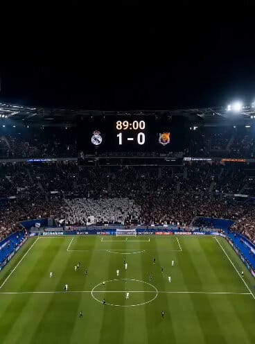 A packed stadium roaring under floodlights, the pitch a vivid emerald green slicked with evenin…