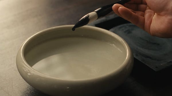 SCENE: Overhead locked close-up, 16:9, on a ceramic water bowl beside a dark inkstone. An adult…