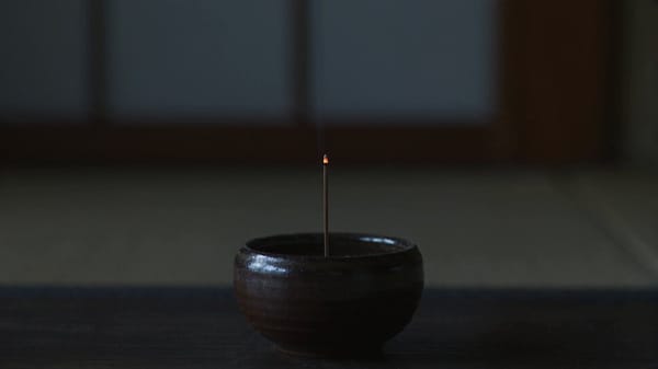 SCENE: Tatami edge at night, 16:9. Locked close-up on a small ceramic incense burner. A single…